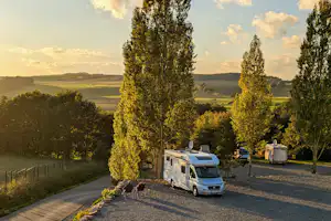 Zwei Wohnmobile auf einem Campingplatzweg, umgeben von hohen Bäumen, mit Blick auf die Landschaft bei Sonnenuntergang.