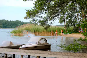 Boot am Steg und Badebereich am Seeufer, Kinder im Wasser, Sandstrand unter Bäumen und Schilf am Ufer.