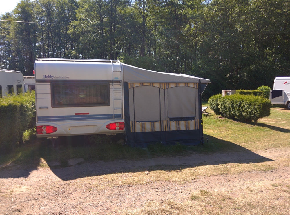 Wohnwagen mit Vorzelt auf einem Campingplatz, gepflasterter Stellplatz und Hecken, im Hintergrund Bäume und ein weiterer Camper.
