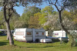 Wohnwagen auf einem Campingplatz im Grünen, mit Picknicktisch und Liegestühlen zwischen Bäumen vor blauem Himmel.