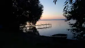 Campingplatz am See bei Sonnenuntergang, dunkle Bäume und ein Steg im Hintergrund, Boot am Ufer.