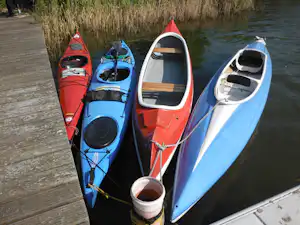 Drei Kajaks liegen nebeneinander am Steg am Wasser, mit Ruderbooten und Schilf im Hintergrund.