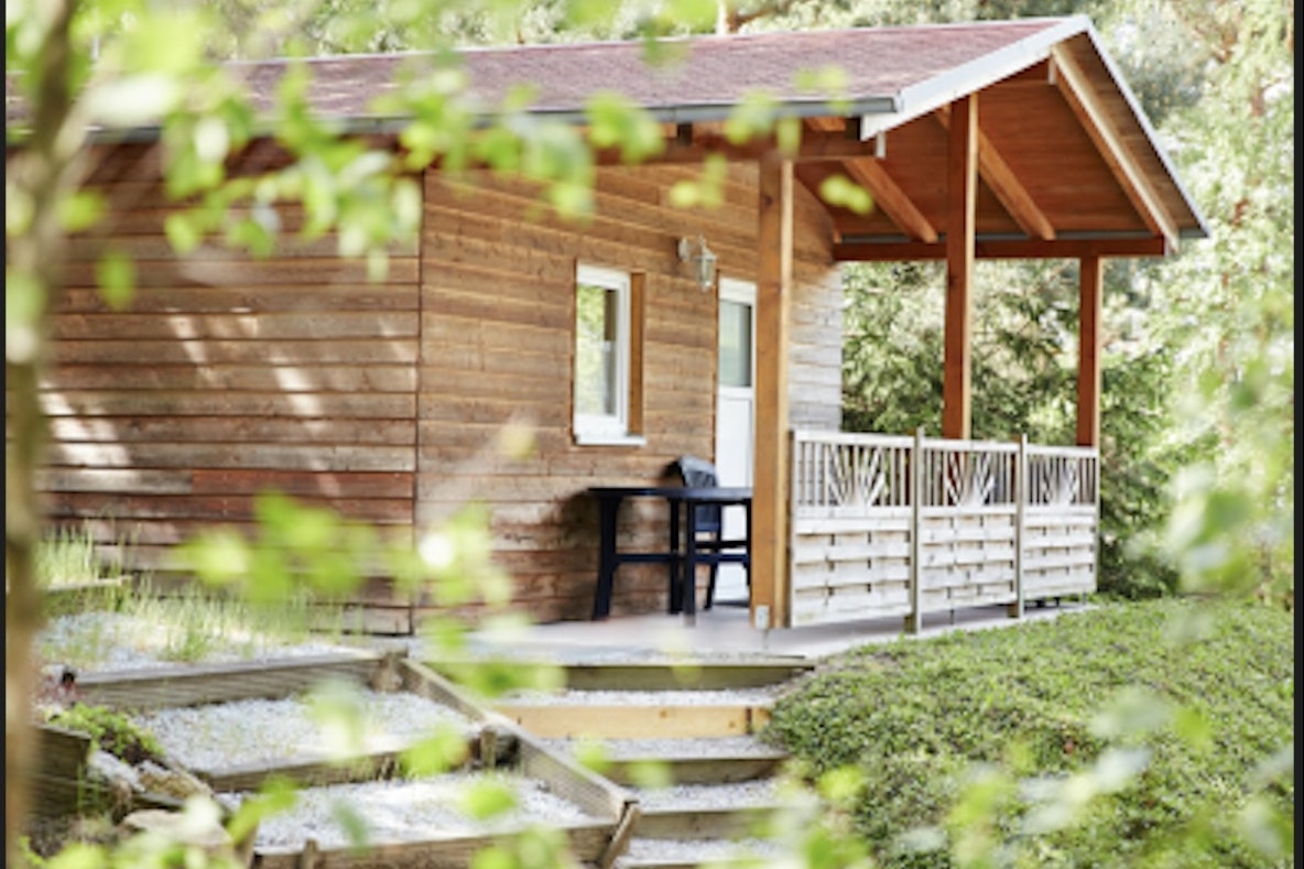Holz-Campinghütte mit überdachter Terrasse, Stühlen und kleinem Tisch, umgeben von Grün und Büschen im Garten.