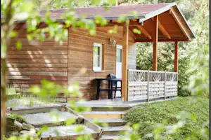 Holz-Campinghütte mit überdachter Terrasse, Stühlen und kleinem Tisch, umgeben von Grün und Büschen im Garten.