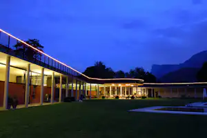 Campinganlage am Abend: beleuchtete Säulen und lange Lichtlinie am Gebäude, Rasenfläche und Pool im Vordergrund.