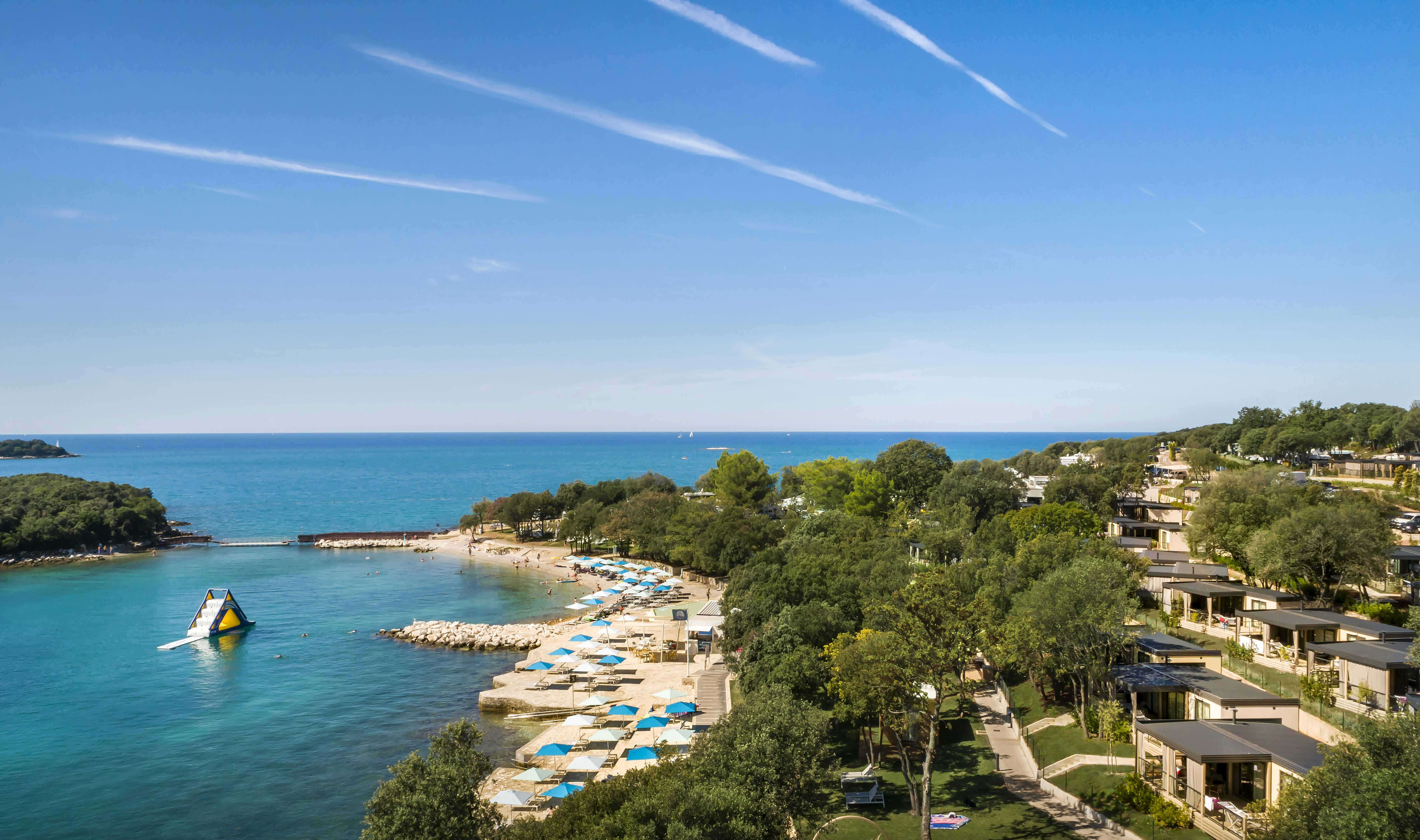 Blick auf eine Küstenbucht mit Strand und Liegen, umgeben von Bäumen und Campingunterkünften am Hang.
