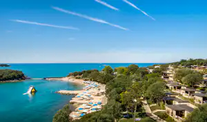 Blick auf eine Küstenbucht mit Strand und Liegen, umgeben von Bäumen und Campingunterkünften am Hang.