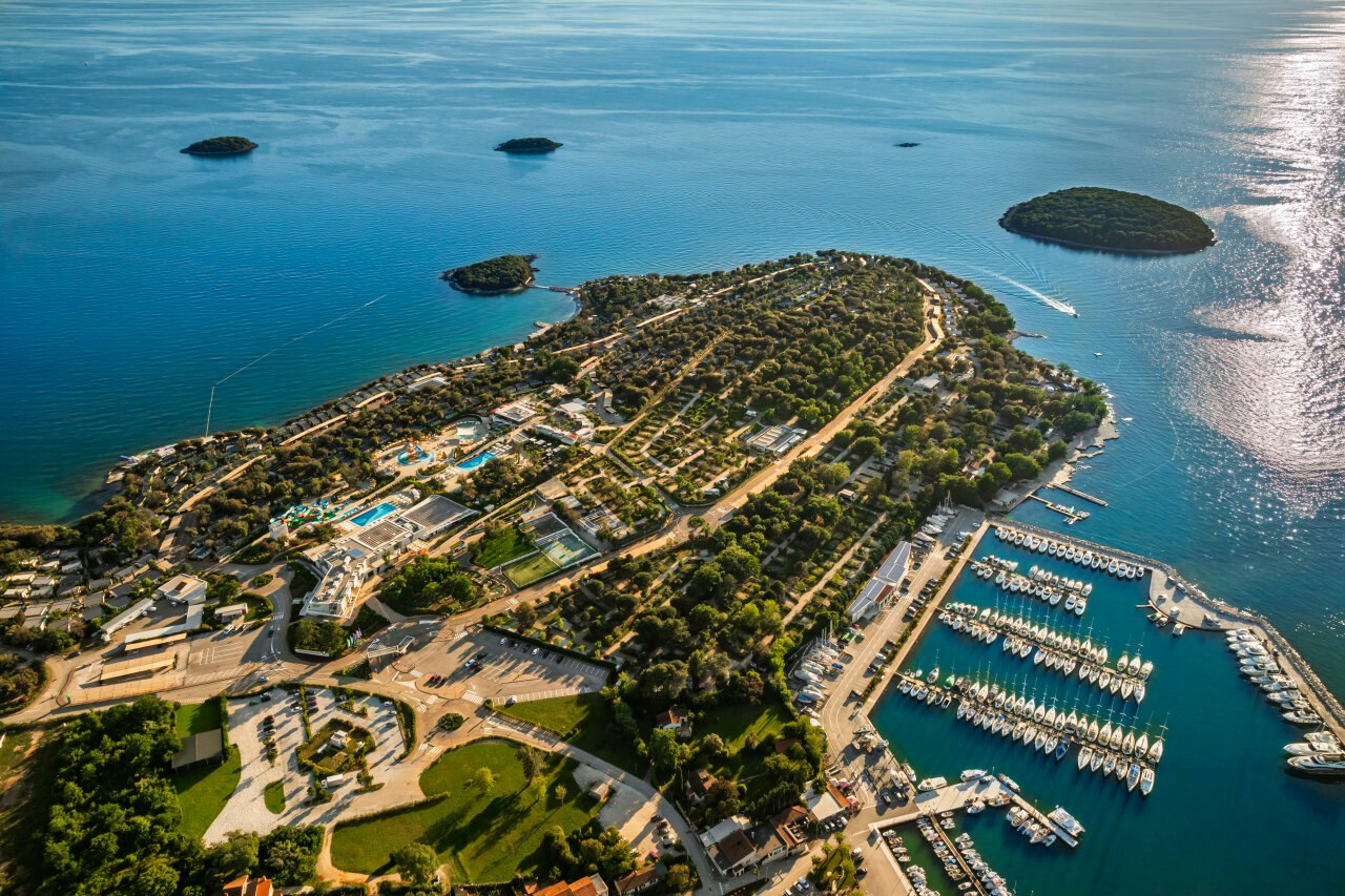 Luftaufnahme eines Campingplatzes an der Küste mit Hafen, Bootsstegen und Stellplätzen zwischen Wegen und Bäumen.