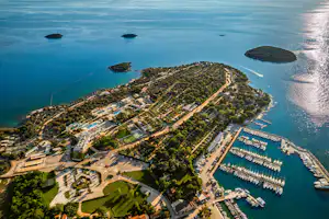 Luftaufnahme eines Campingplatzes an der Küste mit Hafen, Bootsstegen und Stellplätzen zwischen Wegen und Bäumen.