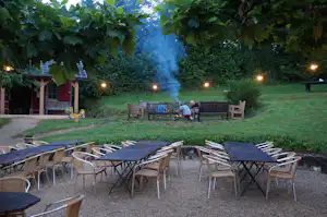Feuerstelle im Campinggarten mit vielen Stühlen und Tischen, beleuchteter Wiese, Rauch und Bäumen im Hintergrund.
