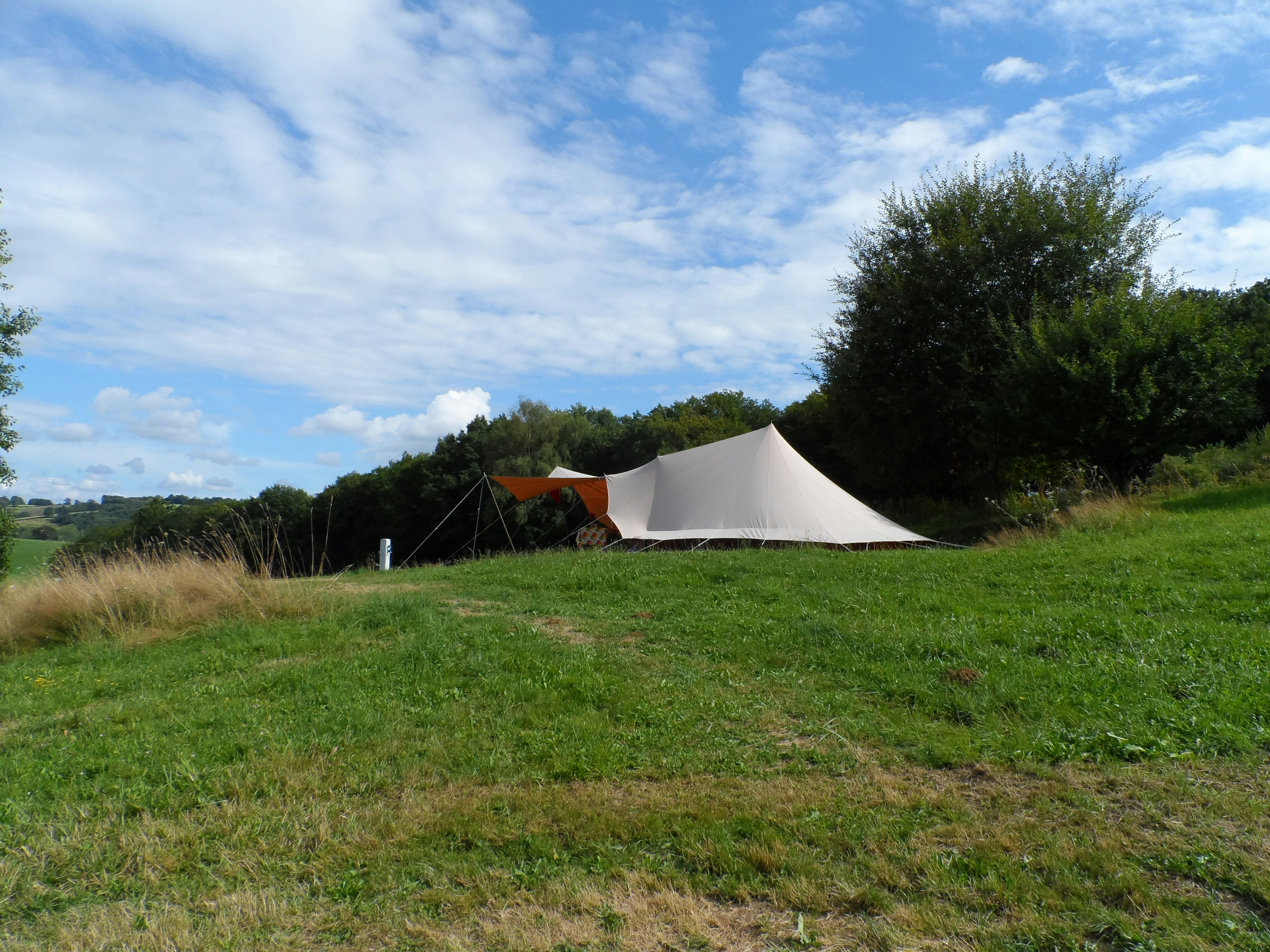 Campingzelt auf grünem Gras am Hang, daneben ein weiteres Zelt, Hecken und Bäume am Rand.