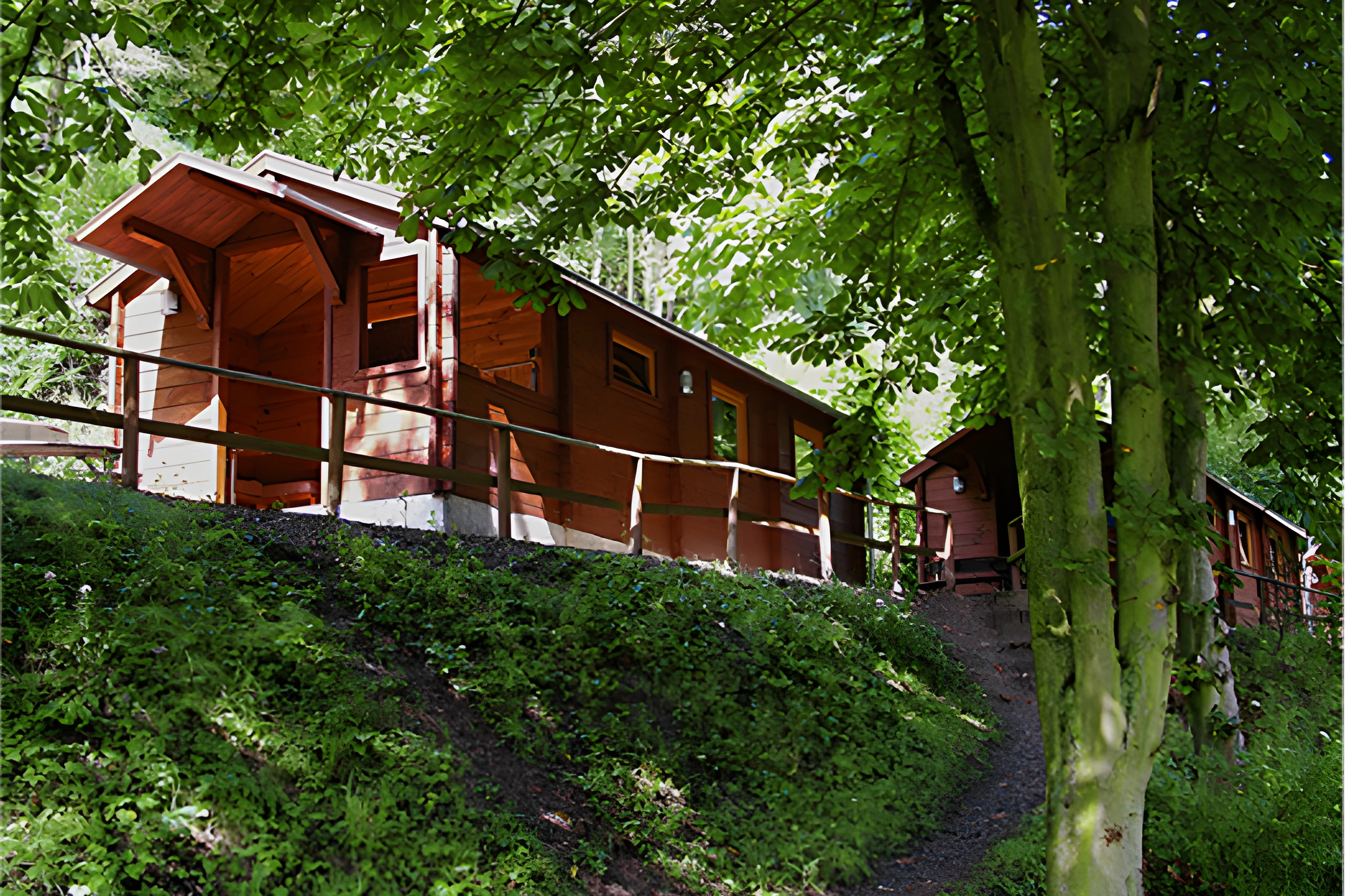 Holzunterkunft mit Balkon und Geländer am Hang, umgeben von Bäumen und einem gepflasterten Weg im Campingbereich.