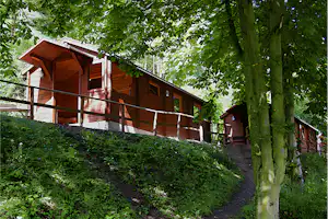 Holzunterkunft mit Balkon und Geländer am Hang, umgeben von Bäumen und einem gepflasterten Weg im Campingbereich.
