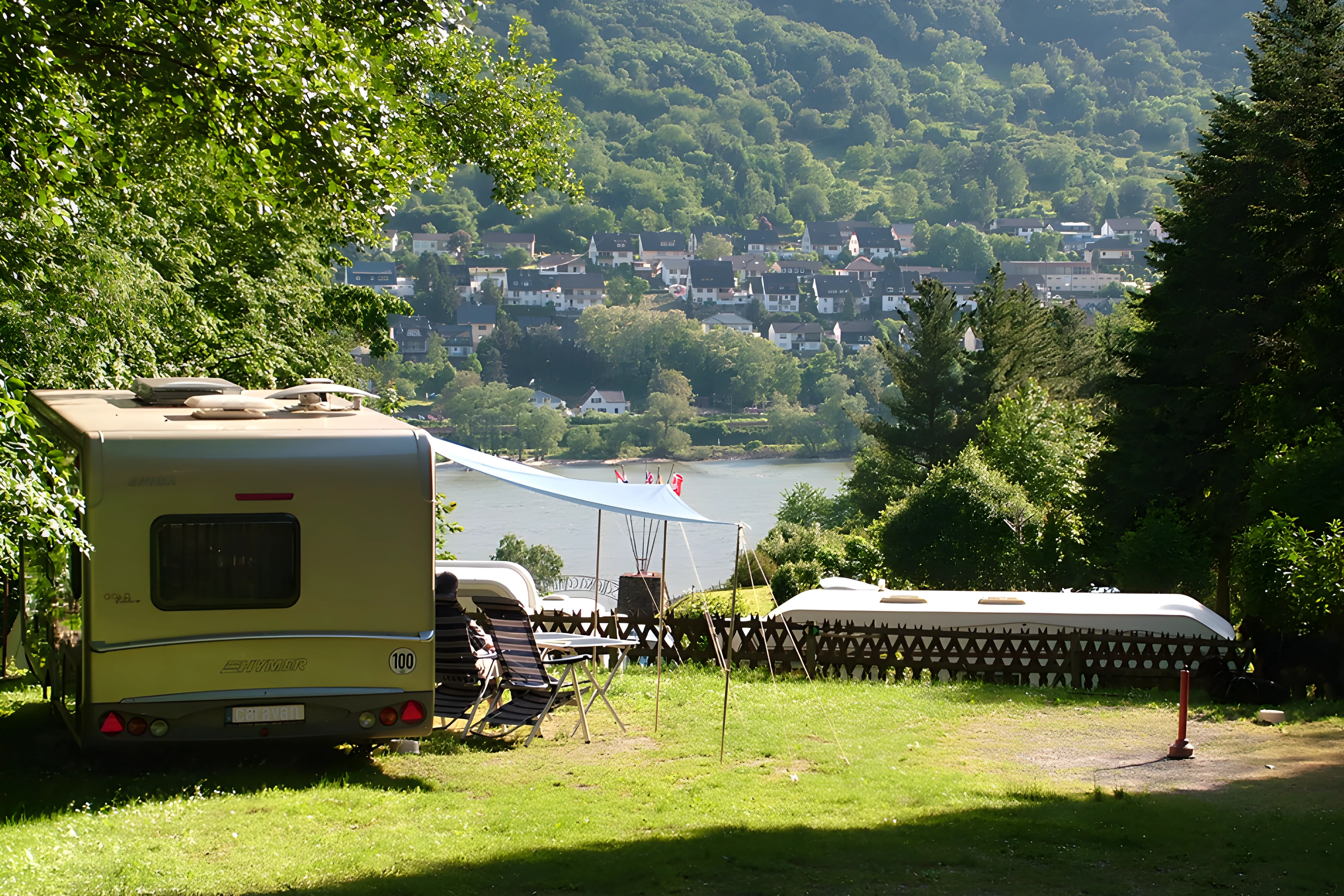 Wohnwagen und Vorzelt auf einem Campingplatz mit Sitzgarnitur, Blick auf den Fluss und die Häuser im Tal.