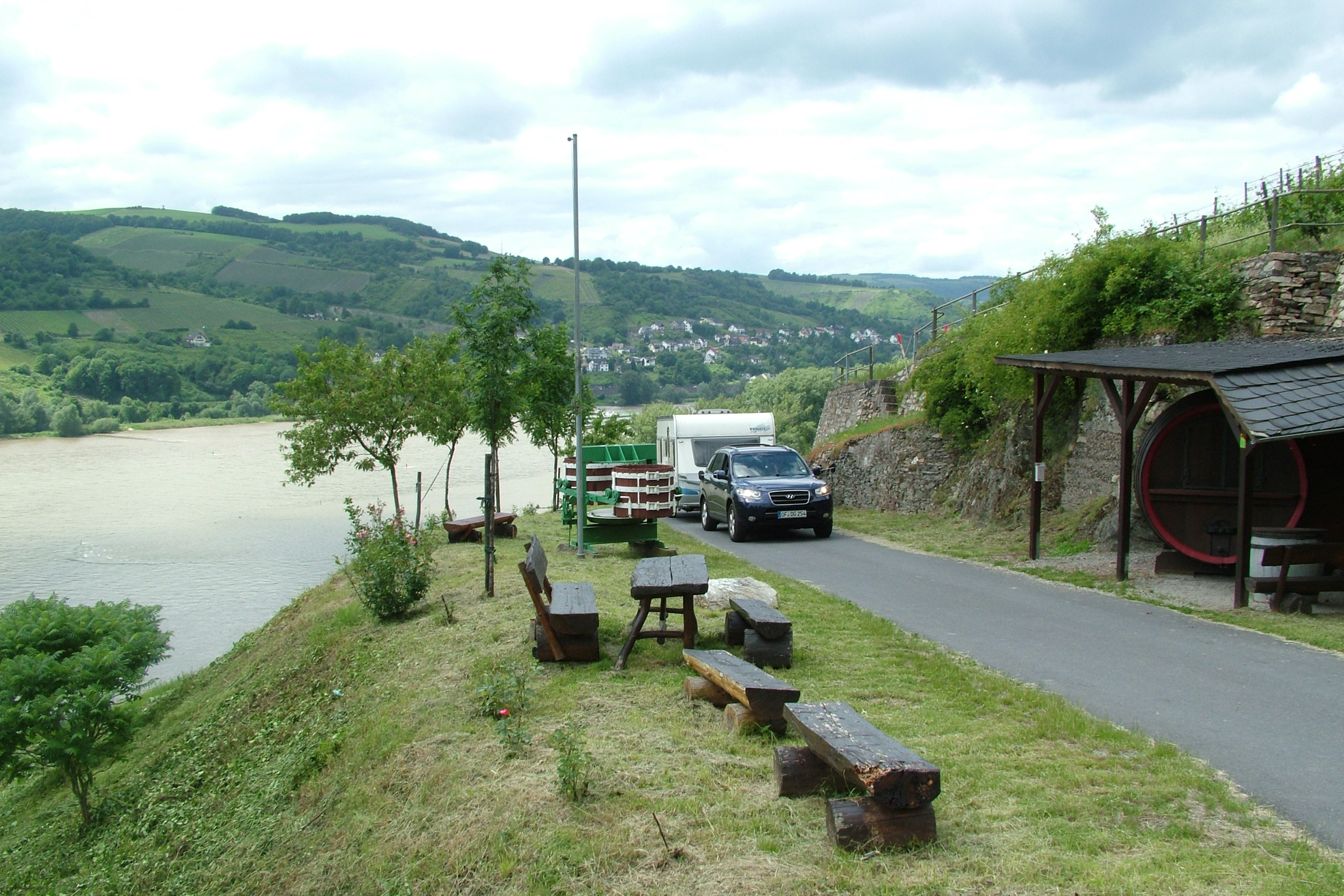 Campingplatz am Fluss: Rasenstreifen mit Bank und Tisch, Stellplätze mit Wohnwagen und Lkw, Straße und Bäume am Hang.