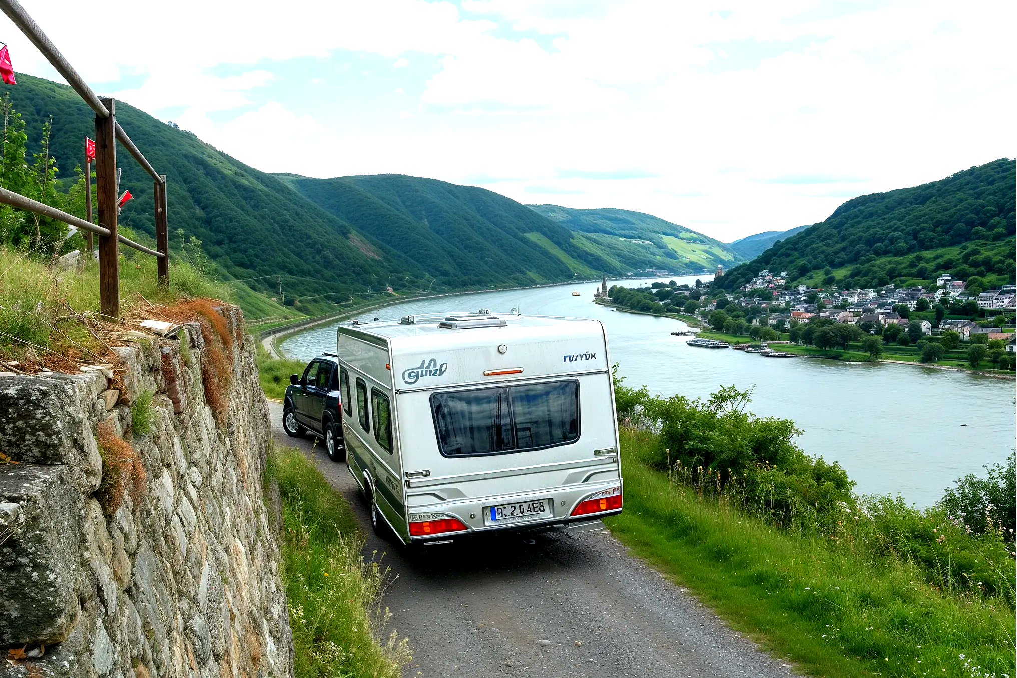 Wohnmobil auf einer schmalen Straße am Fluss, mit Blick auf grüne Hügel und ein Campinggebiet am Ufer.