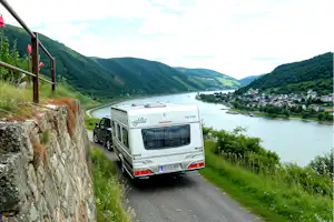 Wohnmobil auf einer schmalen Straße am Fluss, mit Blick auf grüne Hügel und ein Campinggebiet am Ufer.