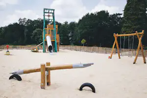 Sandspielplatz am Campingplatz mit Klettergerüst, Rutsche und Wippe, umgeben von Hecken und Bäumen.