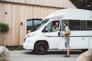 Weißes Wohnmobil am Campingplatz vor einer Holzfassade, zwei Personen stehen am Eingang.