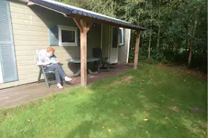 Terrasse am Bungalow mit Holzdach, Tisch und Stühlen, daneben Rasen und Bäume am Rand des Grundstücks.