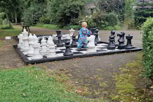 Schachspiel auf einem großen Brett im grünen Campinggarten, mit Figuren und einem Kind im Hintergrund.