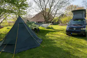 Campingzelt auf einer grünen Wiese, daneben ein Wohnmobil, im Hintergrund Bäume und ein Gebäude mit Satteldach.