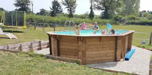 Holz-Whirlpool auf Kiesfläche im Campinggarten, daneben Liegen und ein Zaun mit Spielbereich im Hintergrund.