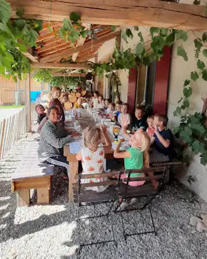 Familie sitzt an einem Holztisch auf dem Campingplatz unter Weinreben, mit Kindern und Erwachsenen im Schatten der Pergola.