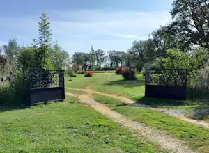Campingplatz-Eingang mit zwei Toren, Schotterweg und Rasenfläche, umgeben von Bäumen und Sträuchern im Grünen.