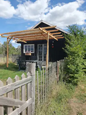 Modernes Holzhaus mit überdachter Terrasse, Gartenmöbeln und großen Fenstern, umgeben von Zaun und Grün.
