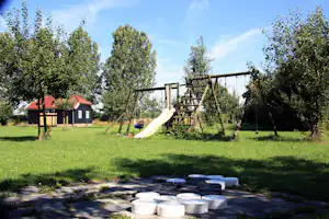 Spielplatz auf dem Campingplatz: Holzschaukel, Rutsche und Wiese mit Gartenhaus im Hintergrund.