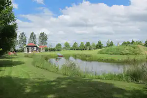 Teich am Campingplatz mit Rasenweg, grünen Bäumen und Wiesen, im Hintergrund ein Gebäude mit rotem Dach.