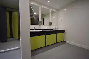 Badezimmer im Campingbereich mit mehreren Waschbecken, Spiegeln und grünen Unterschränken, hellen Deckenleuchten und Fliesenboden.