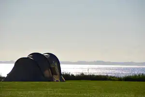Campingzelt am Seeufer auf einer Wiese, im Hintergrund Wasser und entfernte Hügel unter klarem Himmel.