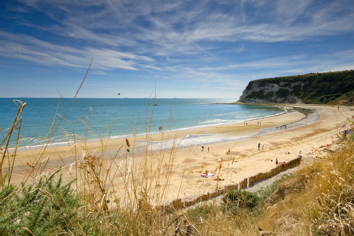 Blick auf einen Sandstrand mit Meer, Kreidefelsen und Spazierweg, im Vordergrund Gräser und am Ufer Menschen.