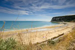 Blick auf einen Sandstrand mit Meer, Kreidefelsen und Spazierweg, im Vordergrund Gräser und am Ufer Menschen.
