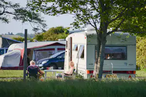 Wohnwagen und Zelt auf einem Campingplatz, davor ein Tisch mit Stühlen und Autos, umgeben von Gras und Bäumen.