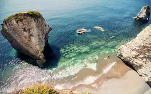 Felsige Bucht mit Sandstrand und klarem Meerwasser, umgeben von großen Felsen und kleinen Wellen.