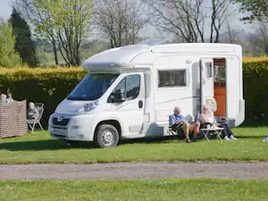 Weißes Wohnmobil auf einem Campingplatz mit Stühlen und Tisch vor dem Fahrzeug, umgeben von Bäumen und Rasen.