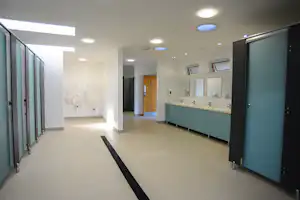 Flur in einem modernen Sanitärgebäude mit hellen Wänden, grauem Boden und mehreren Türen sowie Waschbecken an der rechten Seite.