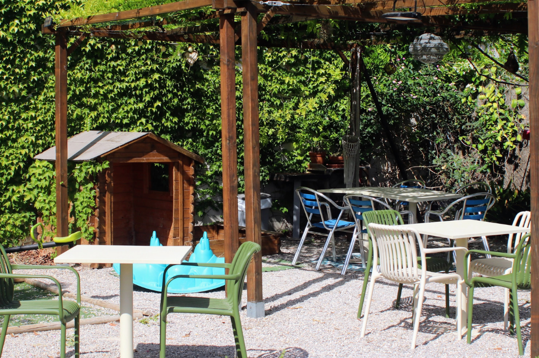 Terrasse mit Tischen und Stühlen unter Holzpergola, daneben ein kleines Holzhaus und grüne Pflanzen im Campinggarten.