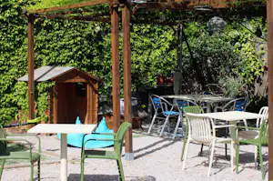 Terrasse mit Tischen und Stühlen unter Holzpergola, daneben ein kleines Holzhaus und grüne Pflanzen im Campinggarten.