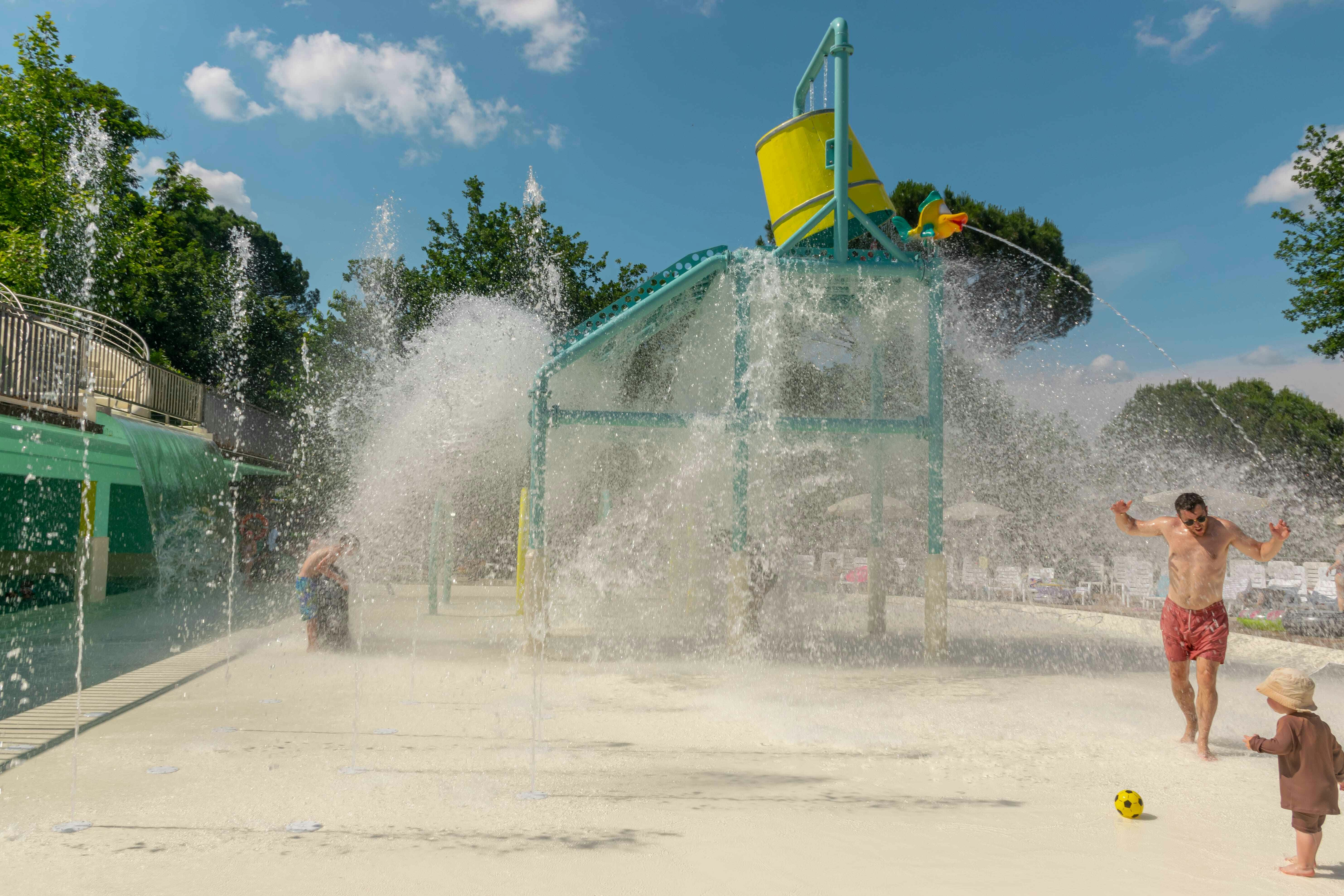 Wasserspielplatz mit großer Sprühfontäne, Kind spielt im Spritzbereich, daneben ein grünes Zelt und Zaun am Rand.