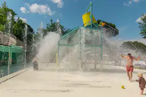 Wasserspielplatz mit großer Sprühfontäne, Kind spielt im Spritzbereich, daneben ein grünes Zelt und Zaun am Rand.