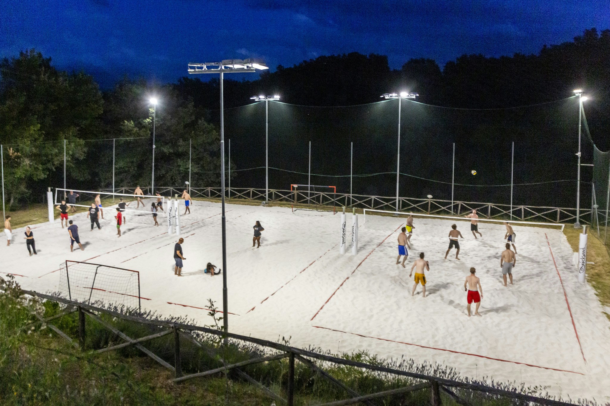 Beleuchteter Beachvolleyballplatz mit Netzen und Linien, mehrere Spieler auf dem Sand, Zäune und Bäume im Hintergrund.