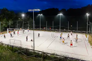 Beleuchteter Beachvolleyballplatz mit Netzen und Linien, mehrere Spieler auf dem Sand, Zäune und Bäume im Hintergrund.