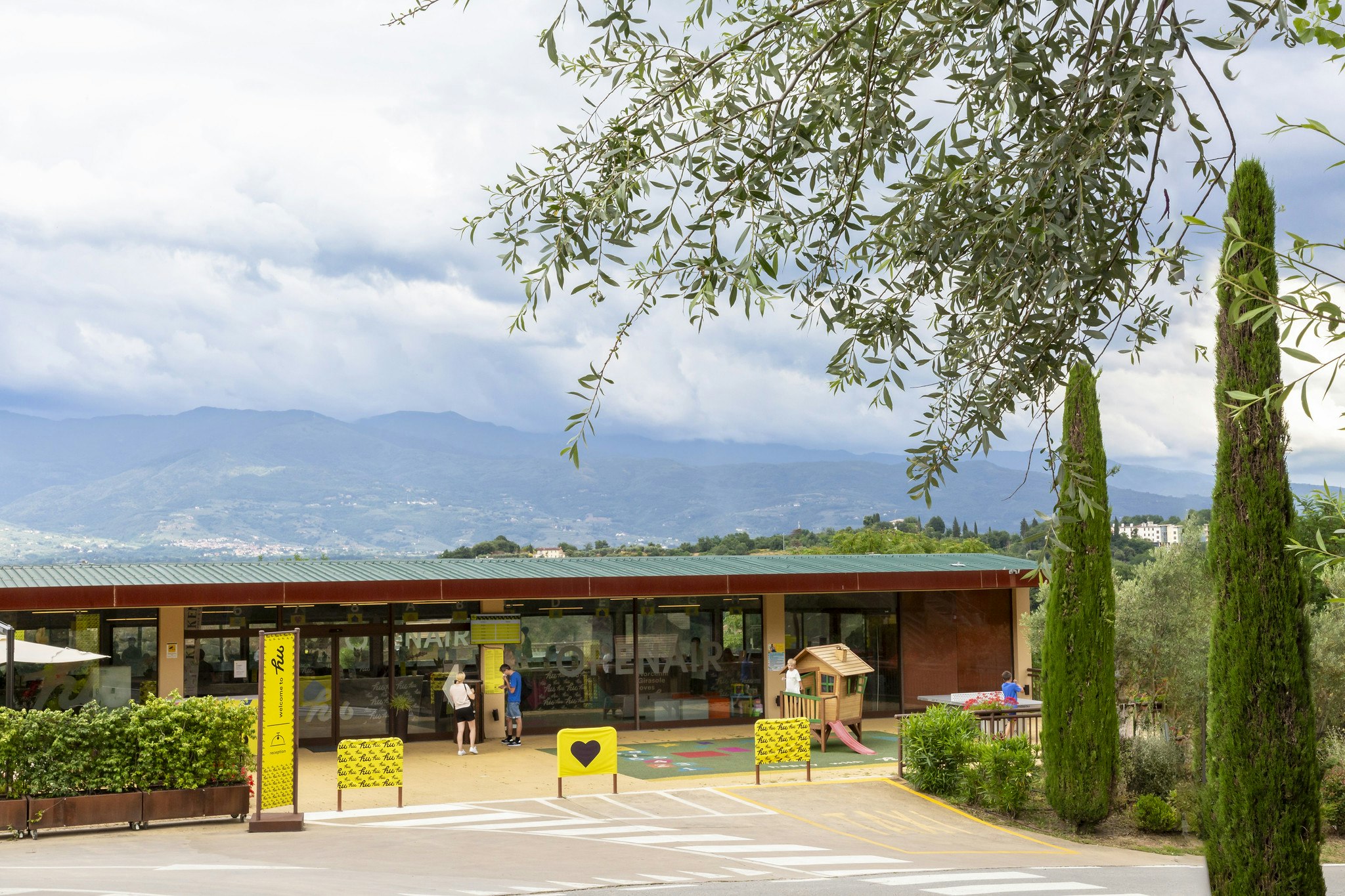 Eingang eines Campingplatzes mit überdachter Rezeption, gelben Sitzbänken und Bäumen, im Hintergrund Berge und Landschaft.