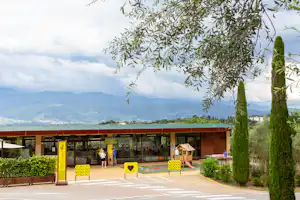 Eingang eines Campingplatzes mit überdachter Rezeption, gelben Sitzbänken und Bäumen, im Hintergrund Berge und Landschaft.