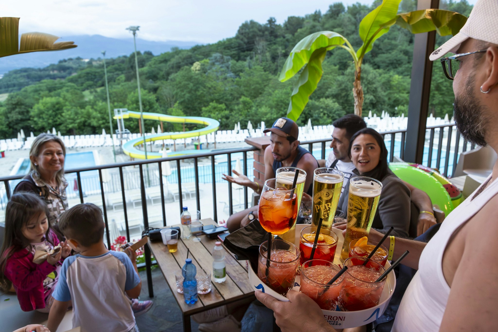 Gruppe am Holztisch auf der Terrasse, Gläser mit Getränken und Kinder schauen zu, im Hintergrund Pool und grüne Hügel.
