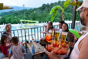 Gruppe am Holztisch auf der Terrasse, Gläser mit Getränken und Kinder schauen zu, im Hintergrund Pool und grüne Hügel.